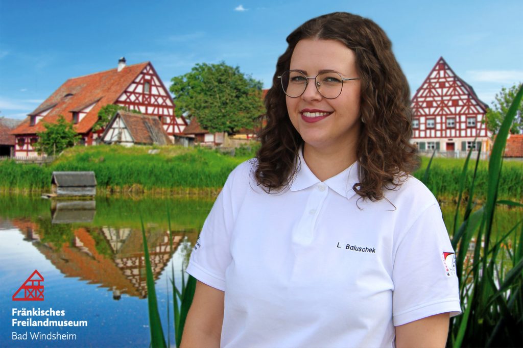 Die Ansprechpartnerin des Fränkischen Freilandmuseums Bad Windsheim, Lisa Baluschek ist im Porträt vor einer unscharfen Ansicht des Museumsbauernhofes und Dorfweihers abgebildet.