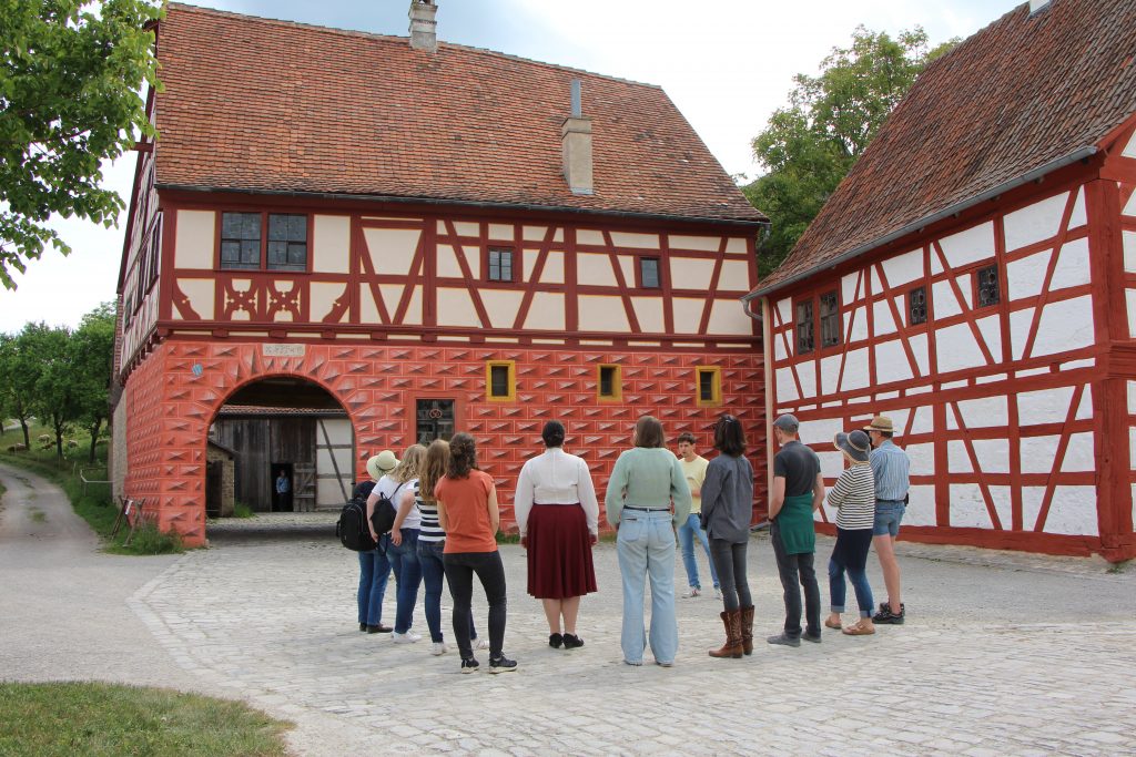 Eine Gruppe Menschen steht auf einem Platz, schaut auf ein großes Fachwerkgebäude und hört einem Rundgangsleiter zu, der vor ihnen steht.
