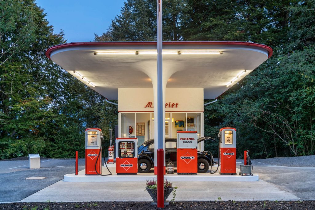 An einer historischen Gasolin-Tankstelle wird ein schwarzer VW-Käfer betankt. Die Tankstelle ist beleuchtet, es herrscht Dämmerung.