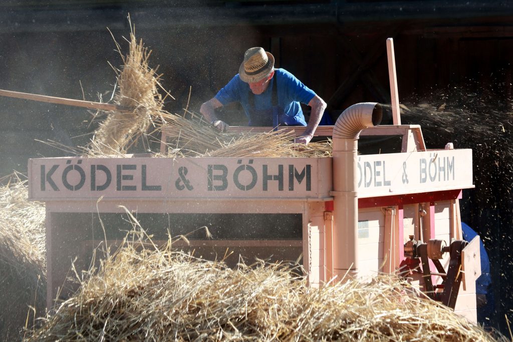 Ein Menn in Arbeitskleidung samt Strohhut arbeitet mit einer historischen Dreschmaschine mit der Firmenaufschrift Ködel & Böhm.