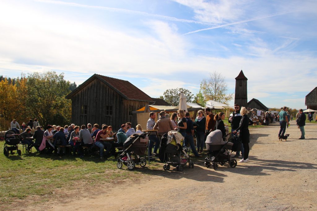 In herbstlicher Atmosphäre sitzen und stehen Menschen an Tischen. Dahinter Marktstände.
