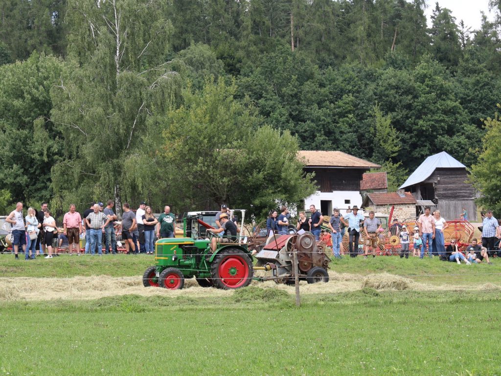 Ein Traktor mit anhängender Heupresse. Drum herum stehen viele Menschen und sehen zu.